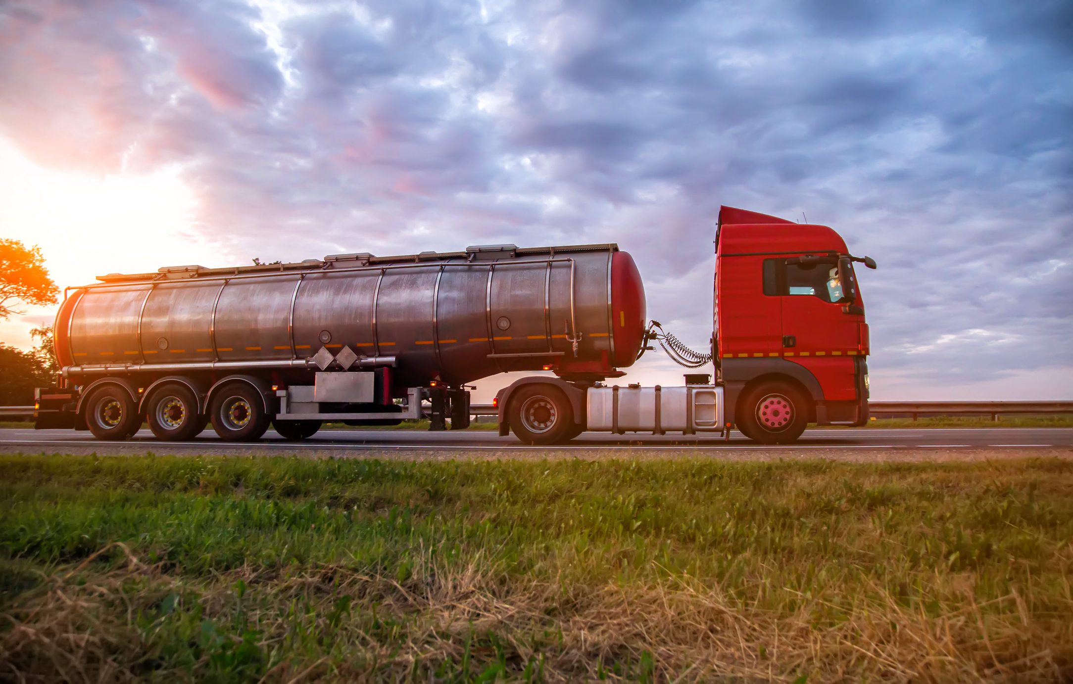 A modern truck with a semi-trailer tanker transports dangerous goods against the backdrop of a sunset in summer. Liquid cargo transportation