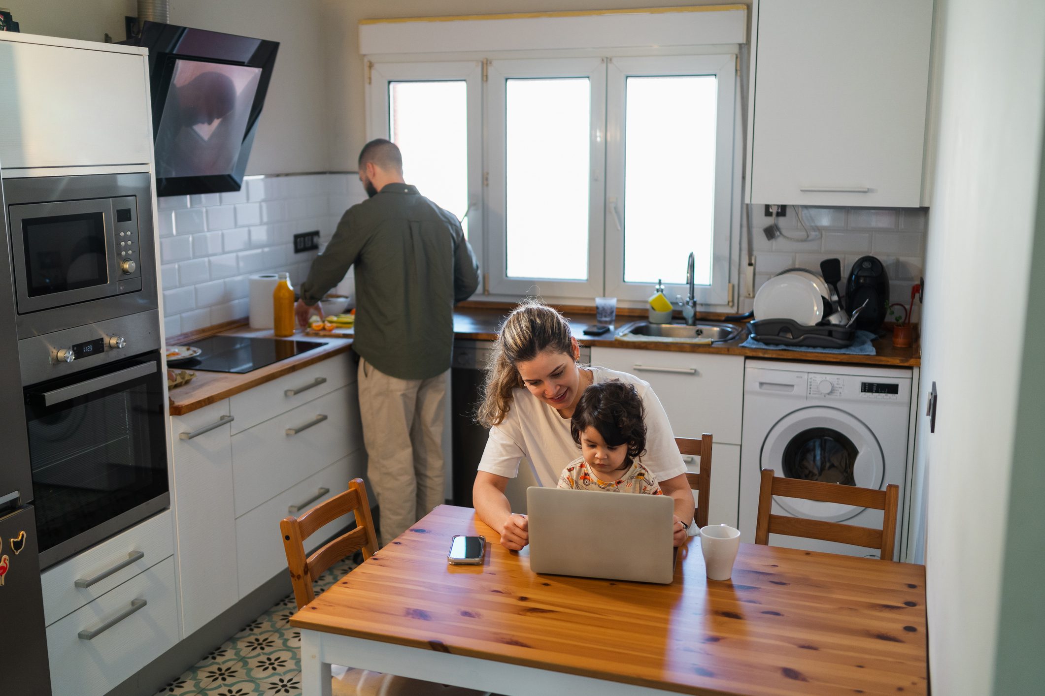 Smiling mother and young child looking at a laptop in the kitchen as father prepares a meal, representing modern family multitasking